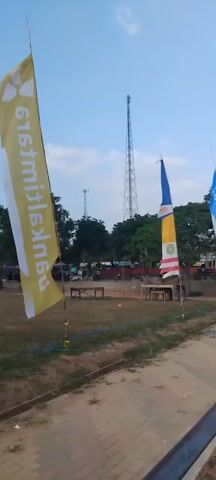 A yellow flag with white text stands in a field. The text is illegible due to the blurry image and its orientation. In the background, trees, a tall transmission tower, a soccer goal, and several people can be seen. The ground appears to be dry grass.