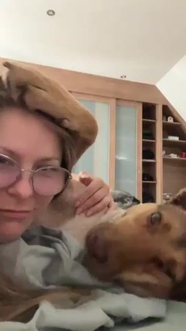 A young woman with glasses lies in bed with her dog. The woman is positioned on the left side of the frame, with her head tilted towards the camera, smiling softly. Her right arm is extended, gently petting her dog. The dog is lying on its back, facing the woman, with its head resting on her chest. Its eyes are closed, and it appears relaxed and content. The dog's tail is held high, curving over the woman's head, almost like a halo.

The setting is a bedroom, suggested by the presence of a bed with green sheets and a gray duvet. Behind the bed, a large wooden wardrobe with sliding doors is visible. The wardrobe has a warm, natural wood finish and appears to have multiple shelves and compartments. The lighting suggests it is daytime, possibly morning or afternoon, as there is a soft, natural light illuminating the scene. The overall mood is one of affection, comfort, and domestic tranquility. The woman's expression and the dog's relaxed posture convey a strong bond between them. There is no visible text in the media file.