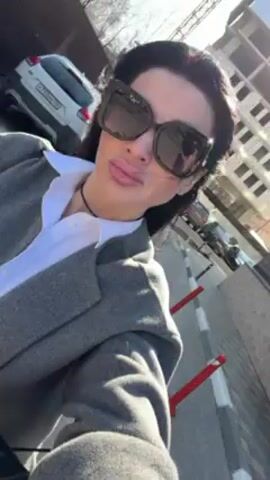 A woman is filming herself on the street, wearing sunglasses, a gray blazer, a white collared shirt, black shorts, and heeled boots. She holds a black shopping bag. She is walking on a paved area with red and white bollards lining the edge. In the background, there are parked cars and a building under construction. The sun is shining.