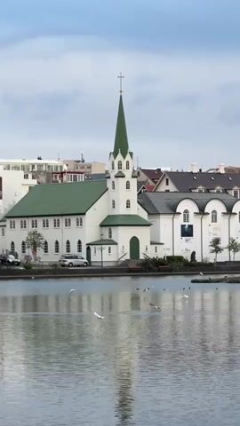 The video shows a white church with a green roof and spire next to a lake. Birds are flying over the water. Several cars are parked on the street.