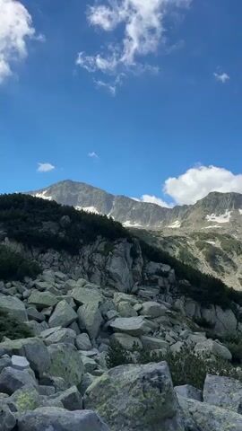 The video pans across a mountainous landscape. In the foreground, there are large, moss-covered rocks and sparse green bushes. The middle ground features a steep, rocky mountainside dotted with patches of dark green vegetation and some remaining snow. In the background, a range of rugged, grey mountains stretches across the horizon, with more snow patches visible on their peaks. The sky is a vibrant blue with scattered white clouds. A turquoise lake is visible nestled in a valley between the mountains in the middle to background of the shot. The scene is illuminated by bright daylight, suggesting it is daytime. There are no people or animals visible in the video.