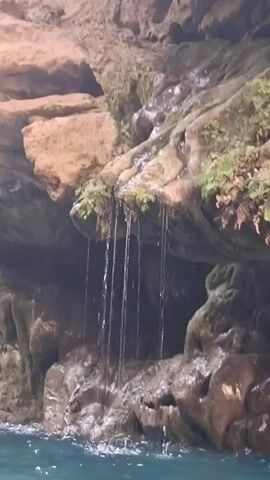 The video shows a close-up view of a natural setting with rocky formations and water. In the foreground, a body of blue water is visible, with ripples on its surface. Above the water, a rock face forms a cave-like structure. From the overhang of this rock face, thin streams of water are cascading down like a small waterfall. Some green moss or vegetation is growing on the rocks where the water is falling. The rocks themselves appear to be layered and eroded, with a sandy or reddish-brown color. The lighting suggests an overcast or shaded environment. The video is taken from a low angle, looking up towards the waterfall and the rock formations.