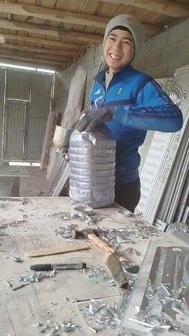 A person in a blue tracksuit and grey beanie stands at a workbench in what appears to be a workshop. The person is holding a large, clear plastic bottle and appears to be manipulating something inside of it with a tool or object. The workbench is cluttered with metal shavings, tools (including a hammer and a marker), and pieces of metal. Behind the workbench are what appear to be decorative metal panels or frames leaning against a wall. The workshop has a wooden ceiling with exposed beams, and a doorway leading to the outside is visible. The lighting in the workshop is dim and the scene appears to be taken indoors.