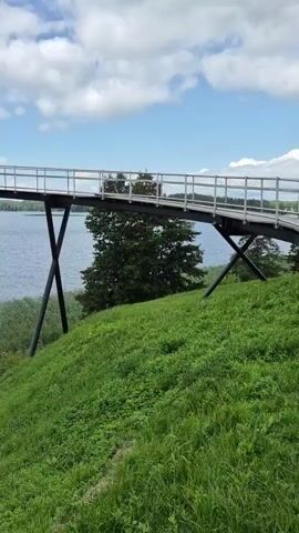 A panoramic view of a curved pedestrian bridge over a body of water. The bridge is supported by two X-shaped black metal structures. People are walking on the bridge. To the right of the bridge, there is a grassy hill. To the left and in front of the bridge is a large lake, bordered by reeds. The sky is blue with scattered white clouds. The camera pans across the scene from right to left.