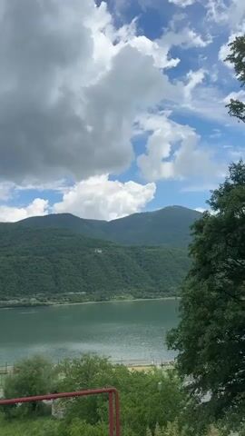 The video captures a serene landscape of a wide, calm body of water, likely a lake or river, flanked by lush, tree-covered mountains under a bright, partly cloudy sky. The water's surface reflects the light, appearing a soft green hue. In the foreground, the perspective shifts slightly to reveal more of the natural environment, with trees framing the lower part of the shot. Along the shoreline, a few structures are visible, including what appears to be a small recreational area with what might be tennis courts or similar sports facilities. The overall impression is one of natural beauty and tranquility, with hints of human presence integrated into the landscape.