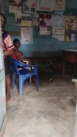A woman opens a doorway and reveals a child sitting in a blue plastic chair. The child, dressed in a red tank top, smiles at the camera. The room has light blue walls covered with posters and papers, a wooden desk, and a bench. The floor is gray concrete. The woman is partially visible, wearing a patterned top and black shorts. Her sandals are visible near the bottom of the door.