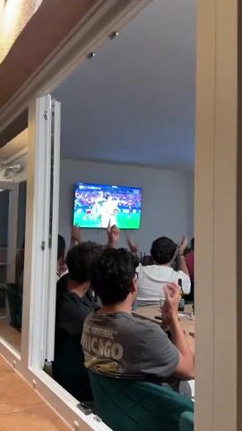 Four people are watching a soccer game on a large television screen mounted on a wall. Two of the people are seen from behind, with their arms raised, appearing to clap or cheer. The other two people are partially visible in the foreground. The television displays a soccer match, with players in white and red jerseys celebrating. The screen shows a score of "POR 0 - 0 ESP" and "90+5". The room appears to be a living room or communal area, with a grey wall behind the television. The lighting is dim, with the primary light source coming from the television screen. The people are seated and facing the television. One person in the foreground is wearing glasses and has dark, curly hair. Another person to their left has lighter brown hair. The focus is on the group watching the game and their reaction to the events on screen.