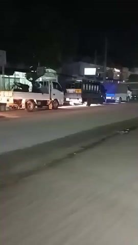 A nighttime street scene shows vehicles moving past, including a white delivery truck and motorcycles. The road appears wet in places, reflecting the lights from shops and buildings lining the street.