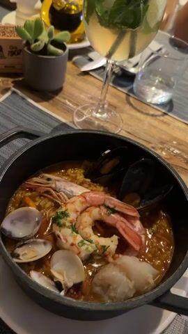The video shows a close-up of a dish of seafood paella in a black pot, garnished with shrimp, mussels, clams, and fish. A glass of mojito with mint and lime sits behind the paella. The scene is set on a wooden table with a striped placemat. In the background, a small potted succulent and a bottle of oil are visible. The video then pans out to reveal a group of people sitting at tables in an outdoor setting, with concrete flooring and various plants. One table has four people seated, with food and drinks. Another table is set with four placemats. The video concludes with an overhead view of the restaurant, showing multiple tables and patrons.