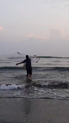 A lone individual, seen from the back, is wading into the ocean. He's carrying a white bag over his shoulder and is dressed in a dark shirt and shorts. The sea stretches out before him under a light sky. Gentle waves break on the shore, and the sand appears dark. The scene is calm and peaceful.
