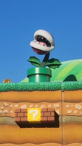 The video shows a stationary, animated Piranha Plant in a green pipe, emerging from a layered dirt structure reminiscent of the Super Mario video game franchise. Below the Piranha Plant, on the dirt structure, is a yellow question mark block encased in brick. The background consists of a clear blue sky and green, rolling hills, also stylized to resemble the Super Mario games. At the bottom of the frame, a sliver of a red and white striped umbrella and the heads of a few people are visible. The Piranha Plant's mouth is open, revealing sharp white teeth. The scene is brightly lit by sunlight.