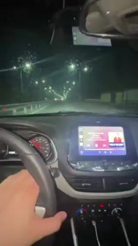 The video depicts a nighttime driving scene from inside a car, likely a late-model Chevrolet, as evidenced by the steering wheel emblem and interior design. Rain is visible on the windshield, with wipers in use. Streetlights illuminate the road ahead, which appears to be a highway or major road based on lane markings and median barriers. The car's infotainment system displays music or a podcast, with an image of a man visible on the screen. The dashboard is visible, showing some of the car's controls and instrumentation. Speedometer needle is visible and moving, indicating the car is in motion.