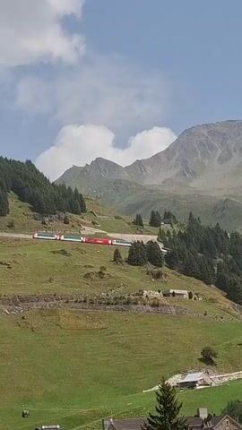 The video shows a mountain landscape with a train and buildings. The train is red and white and is on tracks that wind up a grassy slope. There are trees on the slopes, and a large mountain in the background. In the foreground, there are several buildings that appear to be hotels or apartments. The sky is blue with some clouds. The video is filmed from a high vantage point, looking down on the scene.