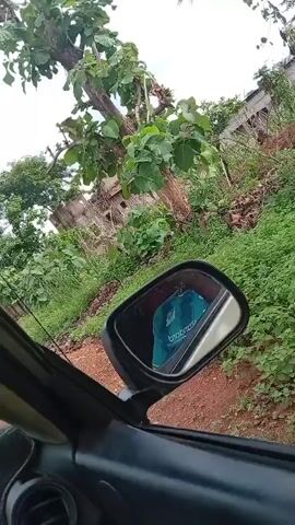 The video shows a view from inside a car driving down a road. There is a motorcycle and a truck driving ahead of the car. The road is paved and there are trees on both sides. The dashboard of the car is visible, with stickers on the windshield. The car is a Toyota RAV4. The side view mirror reflects a green object. The video is shaky.