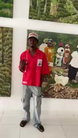 The video shows a man standing in front of paintings in what appears to be an art gallery. He is wearing a red short-sleeved shirt with the word "FORGE" printed on the front, light grey cargo pants, black sandals, and a baseball cap. He poses for the camera, initially making a hand gesture, then touching his face. Behind him are three framed paintings. One painting depicts a forest scene, another shows a group of women in colorful clothing standing near a pile of what looks like cassava. The women are depicted interacting with each other, some holding money and wearing headscarves. The third painting is partially visible and appears to depict another forest scene. The gallery has white walls and tiled flooring. The focus of the video shifts between the man and the painting of the women with cassava.
