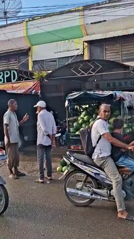 The video depicts a busy outdoor market scene. In the foreground, a black Mitsubishi truck is parked, its bed filled with large sacks of what appear to be onions. In front of the truck, various vegetables are displayed on blue and red tarps, including tomatoes, onions, and leafy greens. A man stands to the right of the truck, facing away from the camera. Further into the scene, two men are conversing near a pile of onions and other produce. More people, motorcycles, and market stalls are visible in the background, with a variety of fruits and vegetables being sold. The environment appears to be a public street with shops and buildings lining the marketplace. The lighting suggests it is daytime.