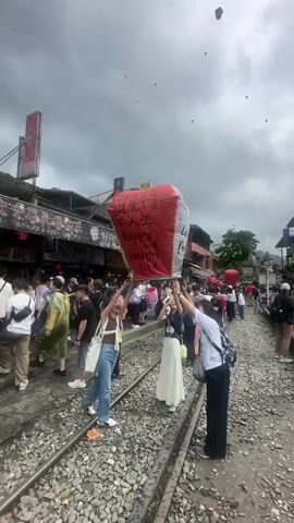 The video shows a crowded street in Taiwan. Many people are standing on and near train tracks. Several people are launching sky lanterns, some of which are red and have writing on them.  The lanterns are floating up into the gray sky.  Many buildings line the street.