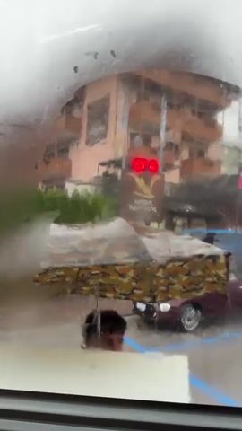The video captures a view through a rain-streaked window, looking out onto a street scene. A multi-story building, appearing to be a hotel or apartment complex, dominates the background. Its facade is a peachy-orange color, with several balconies. In front of the building, a sign for "ADLER NEUEM" is visible. A traffic light displaying red is illuminated.

In the foreground, several cars are parked. One car, a dark red convertible, is prominent in the center. A camouflage tarp covers something in the middle ground. The weather is clearly inclement, with heavy rain distorting the view through the window. Water droplets obscure parts of the scene, and the overall impression is a somewhat hazy and wet atmosphere. The lighting suggests it is daytime, but the overcast and rainy conditions create a muted light. A hand briefly appears at the bottom right corner of the frame, wiping at the window. The scene appears to be in a town, given the presence of buildings, a street, and parked cars. The visible text "ADLER NEUEM" along with the overall style of the building and the setting suggest a location in the Alps, likely a tourist-oriented area given the hotel sign. The emotion conveyed is one of being indoors, observing the weather from a sheltered vantage point.