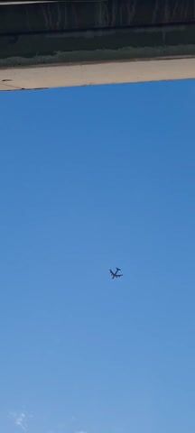 A plane flies across a blue sky with a few sparse white clouds. The plane is dark in color and appears to have four engines. The camera is pointing upwards from the ground.