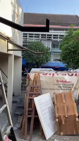 The video pans across an outdoor area filled with assorted items. In the foreground, there are wooden easels, a large framed document or poster leaning against them, and various pieces of wood and metal. Behind these items, large banners or tarps are draped over stacked objects, some bearing text and images. A building with multiple windows and a red-tiled roof is visible in the background, along with trees and a blue tarp canopy. The overall impression is one of clutter, possibly related to an event or storage. No people are clearly visible or engaged in any actions. The environment appears to be daytime, with natural lighting.