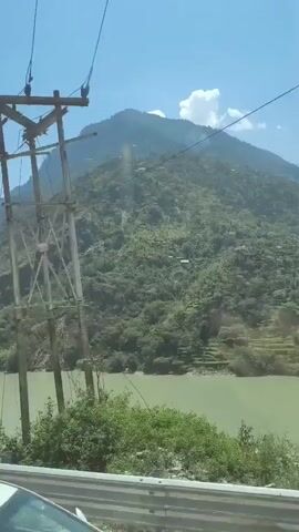 The video shows a view from inside a vehicle looking out a window. In the foreground, a black car window frame and a metal safety barrier are visible. Beyond the barrier, a light green river flows between steep, tree-covered mountains. The sky is clear and blue. A small, black electric fan is mounted above the window, partially obscuring the view. The fan has a white circular logo with the name "SHINA" in black lettering. A black cord hangs from the fan. The vehicle appears to be moving, as indicated by the slight blur of the landscape.