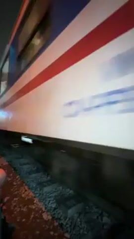 At night, a moving passenger train speeds through a narrow alleyway while several people stand on the side and take photos. The train is painted white and has a thick red stripe running horizontally between two thin blue stripes. The alleyway is dark, but light from inside the train shines through its windows and illuminates the people standing beside the tracks.