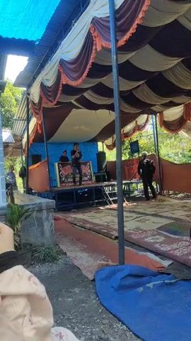 The video shows an outdoor event, likely a gathering or celebration, taking place under a large tent with brown and white fabric draping. In the background, on a raised platform, a man is speaking into a microphone. To the left of the platform, two other individuals are visible. The ground is covered with various mats, including a large blue tarp in the foreground and patterned rugs. Two young children are playing on the mats, one in a plaid shirt and cap, and the other in a pink top. In the distance, there are trees and foliage, indicating an outdoor setting. The overall atmosphere appears to be casual and festive.