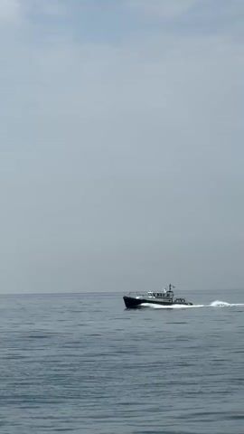 A white and dark blue boat with a white cabin travels across the ocean from left to right. The boat is leaving a white wake behind it. The water is a dark blue and the sky is a pale blue with white clouds. The video is filmed from a stationary position, likely on another boat or the shore.
