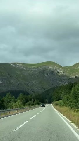 The video shows a car driving on a two-lane road. The road is paved and has white lane markings. On the left side of the road, there is a metal guardrail and a grassy shoulder. On the right side of the road, there is a grassy shoulder and trees. In the background, there are rolling green hills with some visible rock formations. The sky is cloudy. The car in the video is dark in color and is driving away from the camera. The car is in the right lane. The car is the only vehicle visible in the video. The video appears to be taken from inside another vehicle.