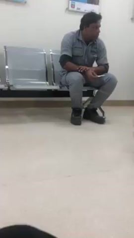 A person in grey coveralls and black shoes sits on a metal bench in a waiting area. The person looks to the right. Another person wearing blue pants and yellow shoes walks by in front of the camera. The walls are white and there is a poster on the wall behind the person on the bench. The floor is a speckled off-white.