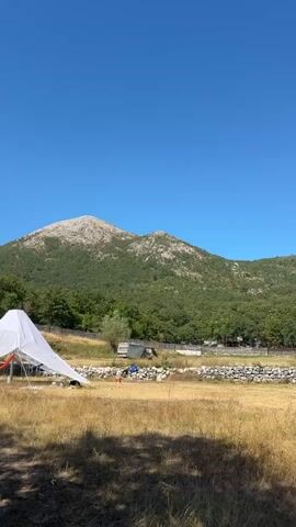 A drone shot pans across a wide, dry, grassy field under a clear blue sky. In the foreground, a large, white, pyramid-shaped tent with its sides pulled back is set up, revealing a red hammock hanging inside. To the right of the tent, a few people are visible near a white camper van, with a low stone wall behind them. In the background, a dense, green forest covers the slopes of a large, rocky mountain that dominates the horizon. The overall impression is of a remote, possibly a campsite, surrounded by nature.