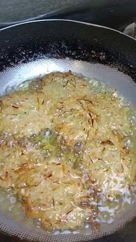 The video shows a close-up, overhead view of several fritters being fried in a pan. The fritters are golden brown and appear to be made of shredded vegetables, possibly zucchini and onions. They are submerged in hot oil, which is bubbling around them. The pan is black and appears to be well-used, with some oil splatters on the rim. The background is blurred but suggests a kitchen environment. The footage is handheld and slightly shaky, creating a sense of immediacy.
