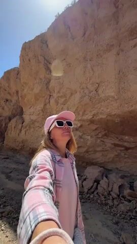 A woman is walking through a dry, rocky canyon under a clear blue sky. She is holding a camera or phone, filming herself in a selfie-style video. She wears a pink baseball cap, white rectangular sunglasses, a pink t-shirt, and a pink and white plaid button-up shirt worn open. Her long hair is light brown.

The canyon walls are tall, steep, and composed of light brown, rocky terrain, appearing very dry and arid. The ground is a dirt and gravel path. The woman looks around the environment, turning her head from right to left, observing the canyon. She has a slight smile at one point, suggesting she is enjoying the exploration. The bright sun indicates it is daytime, likely mid-day, with no clouds in sight. The scene is consistent with the provided location context of Ak Say, Kyrgyzstan.
