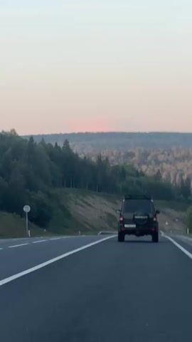 The video shows a black SUV driving on a paved road. The road has white lines marking the lanes. On the left side of the road, there is a signpost with a white circular sign. The landscape features rolling hills covered in trees. The sky is a pale pinkish-orange, suggesting either sunrise or sunset. The SUV is in the right lane, moving away from the camera. There are some other cars visible in the distance on the road. The overall scene is one of a scenic drive through a natural landscape.