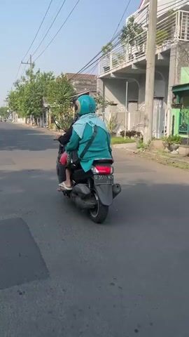 Two individuals are riding on a scooter on a paved road. The person in the back is wearing a light blue helmet and a turquoise jacket. The person in the front is wearing dark clothing. The scooter appears to be dark in color. They are traveling away from the camera. The environment is a street with houses and trees visible on the sides. The sky is clear and the lighting suggests it is daytime, possibly morning or afternoon.
