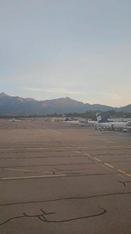 The video shows an airport tarmac with several airplanes parked. The sky is clear and a mountain range is visible in the background. The foreground shows the tarmac with yellow and white markings. Several airplanes with the livery "AIR CORSICA" are visible. Some ground equipment and vehicles are also present around the airplanes. The lighting suggests it is either early morning or late afternoon.