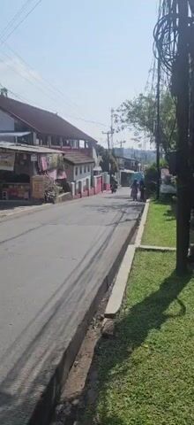 The video captures a street scene with a paved road lined with grass and a sidewalk. On the left side of the road, there are buildings with red roofs and pink walls, some with shops. Several people are visible, some on motorcycles and others walking. Trees and utility poles are also present in the environment. The sky is clear and the sun is shining, casting shadows on the road.