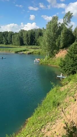This video showcases a scenic lake on a bright, sunny day. The water is a vibrant turquoise, reflecting the clear blue sky dotted with fluffy white clouds. Lush green trees and foliage frame the lake, creating a tranquil natural setting. Several individuals are enjoying various water activities, including paddleboarding and kayaking. One person is seen paddleboarding near the shore, while others are further out on the lake in kayaks. The scene suggests a leisurely day spent outdoors, with the participants appearing relaxed and engaged in their chosen activities. The time of day is likely midday or early afternoon, given the strong sunlight and clear skies. The overall atmosphere is one of peaceful recreation in a beautiful natural environment.