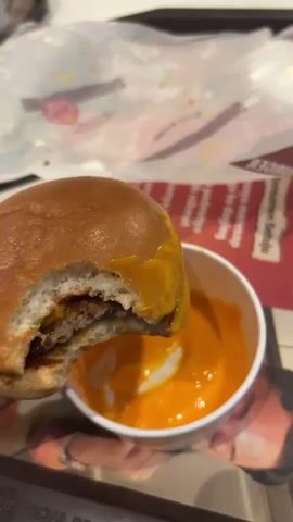 The video shows a partially eaten hamburger dipped in a bowl of orange sauce. The hamburger bun is shiny and brown. The hamburger has a meat patty and a slice of cheese. The orange sauce is in a small white bowl and has a swirl of white sauce mixed in. The background includes a McDonald's tray with a promotional advertisement on it and a crumpled piece of plastic wrap.