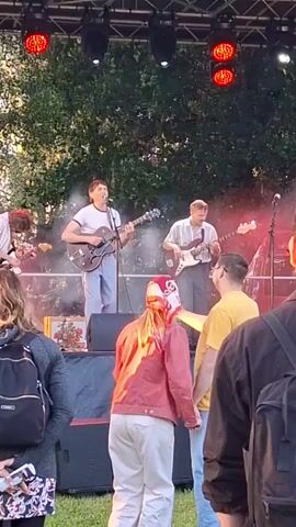 A live outdoor concert is taking place on a stage set up on grass. Four male musicians are performing on stage. The lead singer, wearing a white t-shirt and light-colored pants, is playing a brown electric guitar and singing into a microphone. To his right, another male musician, in a light-colored striped shirt and dark pants, plays a white bass guitar. A third musician, also in a light-colored striped shirt and dark pants, plays a guitar on the left side of the stage, partially obscured. A fourth musician is also visible on the right side of the stage, playing an instrument. Red stage lights are visible above the performers, and some smoke or haze is present in the air. In front of the stage, a crowd of people is watching the performance. The focus is on a woman in the foreground wearing a pink jacket and white pants, with a red and white patterned scarf around her head. She is facing the stage, with her back to the camera. To her right, a man in a yellow t-shirt and blue jeans is also facing the stage. Several other audience members are visible in the foreground and background, some with backpacks. The overall environment is outdoors with trees visible behind the stage, suggesting a park or open-air venue. The lighting suggests it is either late afternoon or early evening.
