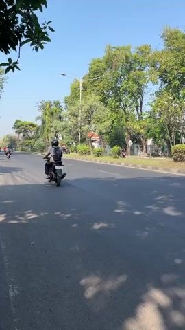 The media file depicts a daytime scene on a paved road in Gelam, Indonesia. The primary subject is a person riding a motorcycle from left to right across the frame. The rider is wearing a helmet and a backpack. The road is bordered by a sidewalk and a median strip lined with trees and bushes. In the background, more trees and some buildings are visible under a clear blue sky, suggesting fair weather. The time of day appears to be late morning or early afternoon, given the bright sunlight and the shadows cast on the road. The overall mood is peaceful and ordinary, capturing a moment of everyday travel. No visible text is present in the media file.