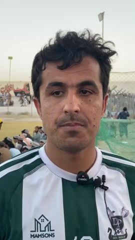 A close-up shot shows a man in a green and white striped athletic jersey. He has dark, curly hair and is looking directly at the camera with a neutral expression. A small black microphone with the logo "candc" is attached to his jersey. In the background, a crowd of people are visible, some sitting on the ground and others standing. A yellow flag on a pole is also visible in the distance. The environment appears to be outdoors, possibly a sports field or stadium, with a fence and netting in the background. The overall lighting suggests it is daytime.