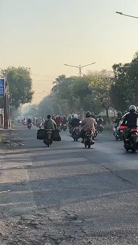 A busy road is filled with a large group of motorcyclists riding in the same direction. The road is paved and has some cracks and debris on the sides. Trees line the road, and overhead power lines are visible. The sky is hazy and the sun appears to be low in the sky, possibly indicating early morning or late afternoon. Most of the motorcyclists are wearing helmets, and some have backpacks or other items secured to their bikes. The overall impression is one of a significant number of people commuting or traveling together on motorcycles in an urban or semi-urban environment.