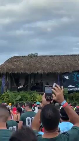 The video shows a crowd gathered outdoors, facing a thatched-roof stage where a band is performing. Many people in the crowd are wearing shirts with the number "00" on the back. Some individuals are holding up their phones, presumably to record the performance. The stage has lush greenery, and two flags flanking the stage each bear a logo of a lion. The sky above is overcast with grey clouds.