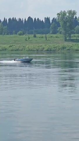 A motorboat with two occupants travels down a wide river, leaving a white wake. The river is calm with gentle ripples. On the far bank, a green, grassy slope is lined with trees. The sky is cloudy. The scene appears to be during daylight.