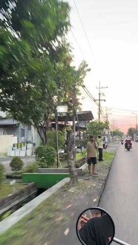 The video captures a street scene in Sumorame, East Java, during what appears to be sunset, indicated by the warm, orange glow in the sky. The camera is mounted on a moving vehicle, likely a motorcycle given the reflection of a hand and handlebars in the lower right corner.

In the foreground, a person in a light brown t-shirt and dark shorts is standing on a sidewalk, holding a phone to their ear. Behind them, another person in a light yellow shirt and dark pants is also on the sidewalk, seemingly interacting with the environment. Further down the street, two individuals are visible on a motorcycle, with one wearing a pink garment and the other darker clothing. The scene is lined with trees and vegetation, with a small ditch or channel running alongside the sidewalk. In the background, buildings and shops are present, and utility poles with numerous wires crisscross the sky.

The overall mood is casual and observational, as the camera moves along the road. The time of day is evident from the soft lighting and the colors in the sky. The location is confirmed to be Sumorame, East Java. No discernible text is legible within the video frames.