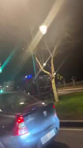 Here is a description of the video:

The video shows a dark, nighttime street scene. A gray car, with license plate visible, is in the foreground. To the side of the car is a black van with red accents parked on a patch of grass next to the road. The van has a light bar on top. Street lights illuminate the scene. A traffic light cycles through green and red signals. The background includes sparse trees and other streetlights lining the road.