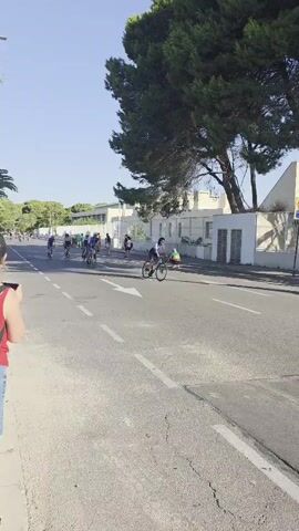 A group of cyclists are riding down a street lined with trees and buildings. The sun is shining and the sky is clear. The cyclists are all wearing helmets and cycling gear. Some of the cyclists are in a race, while others are just enjoying a ride. A pedestrian is visible in the background. There are cars parked on the side of the road and traffic signs. The road has white lane markings.