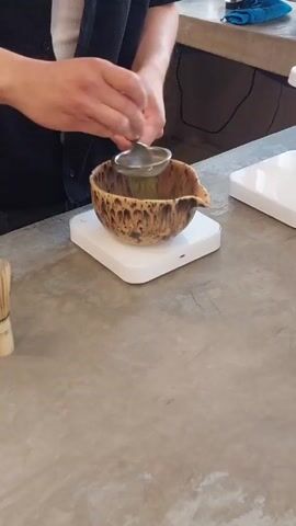 Here's a description of the video:

A person is shown preparing matcha in a ceramic bowl. They are using a small, handled sieve to sift a bright green powder into the bowl. The bowl sits on a small, white, square scale. To the left of the bowl is a traditional bamboo whisk. The countertop is a neutral gray. The person is wearing a black vest over a white shirt.