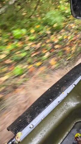 The video shows a first-person perspective of a vehicle driving through shallow, muddy water in an outdoor, wooded environment. The water is brown and disturbed, indicating movement. The vehicle appears to be an off-road vehicle, possibly an ATV, with a tire and part of the vehicle's frame visible in the foreground. Green foliage and fallen autumn leaves are visible along the sides of the path. The camera is shaking, suggesting the vehicle is in motion over uneven terrain.
